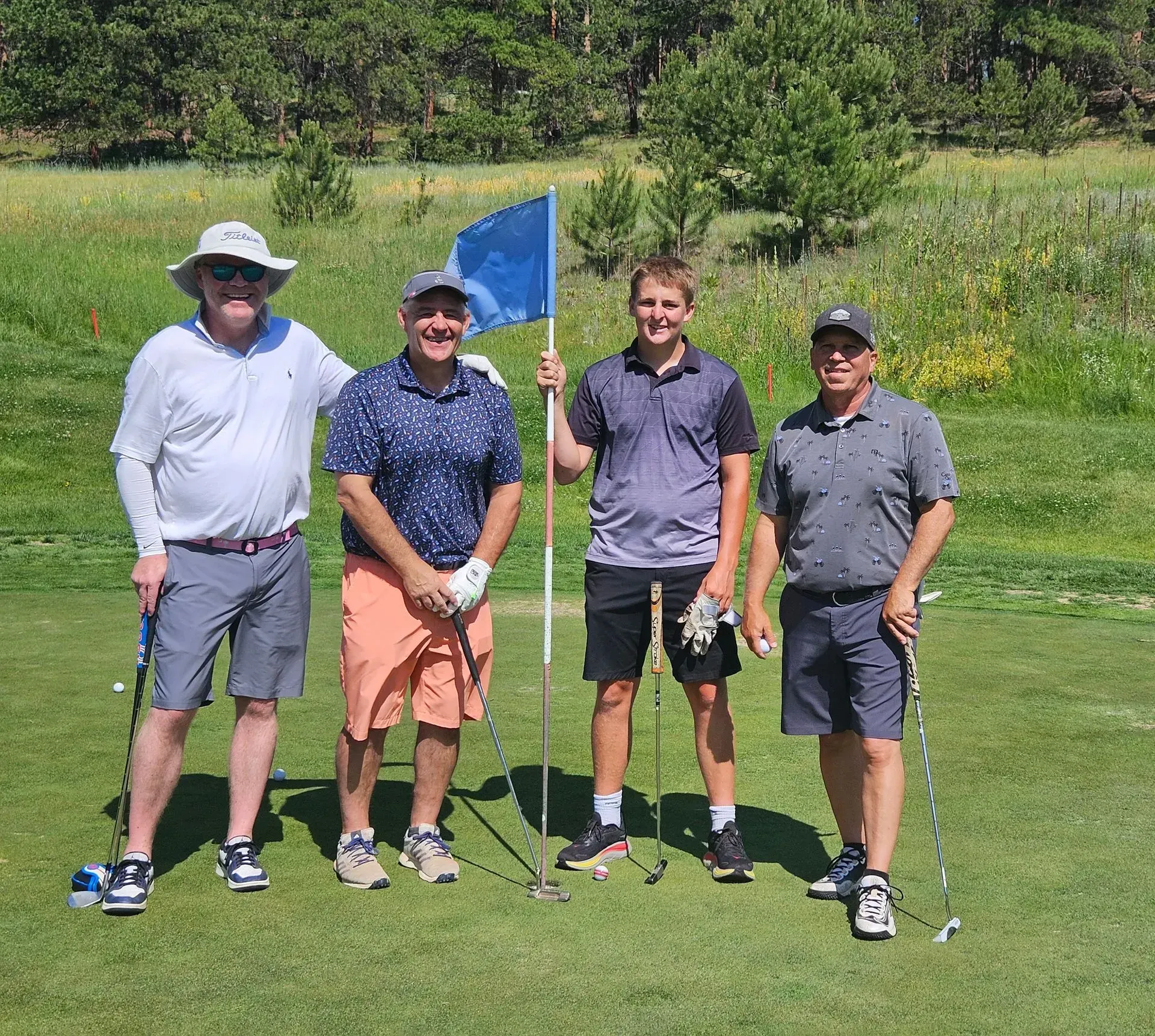 Four men on a golf course posing with a flag. They are smiling, wearing golf attire, and are in a sunny field.