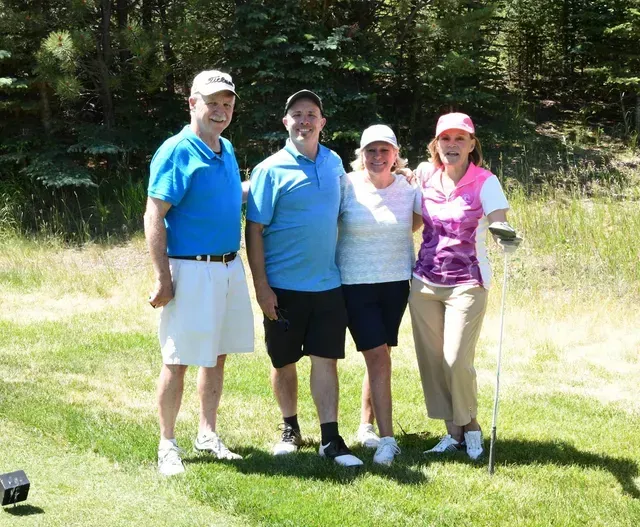 Four people smiling, posing on a grassy golf course. Two men and two women in golf attire.