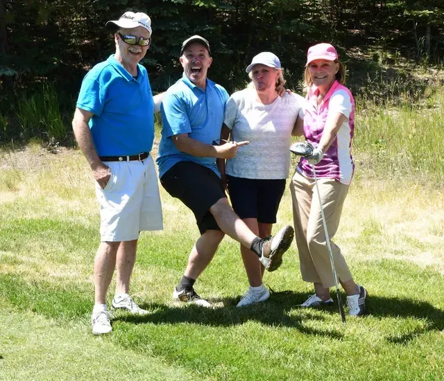 Four people, including a golfer, pose cheerfully on a golf course. The man points, smiling.