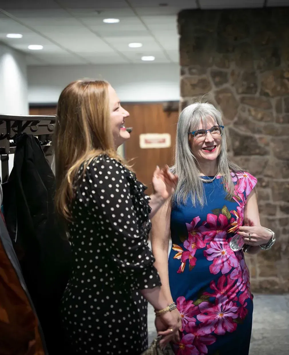 Two women laughing in a hallway, one in a polka dot dress, the other in a floral dress, near a stone wall.