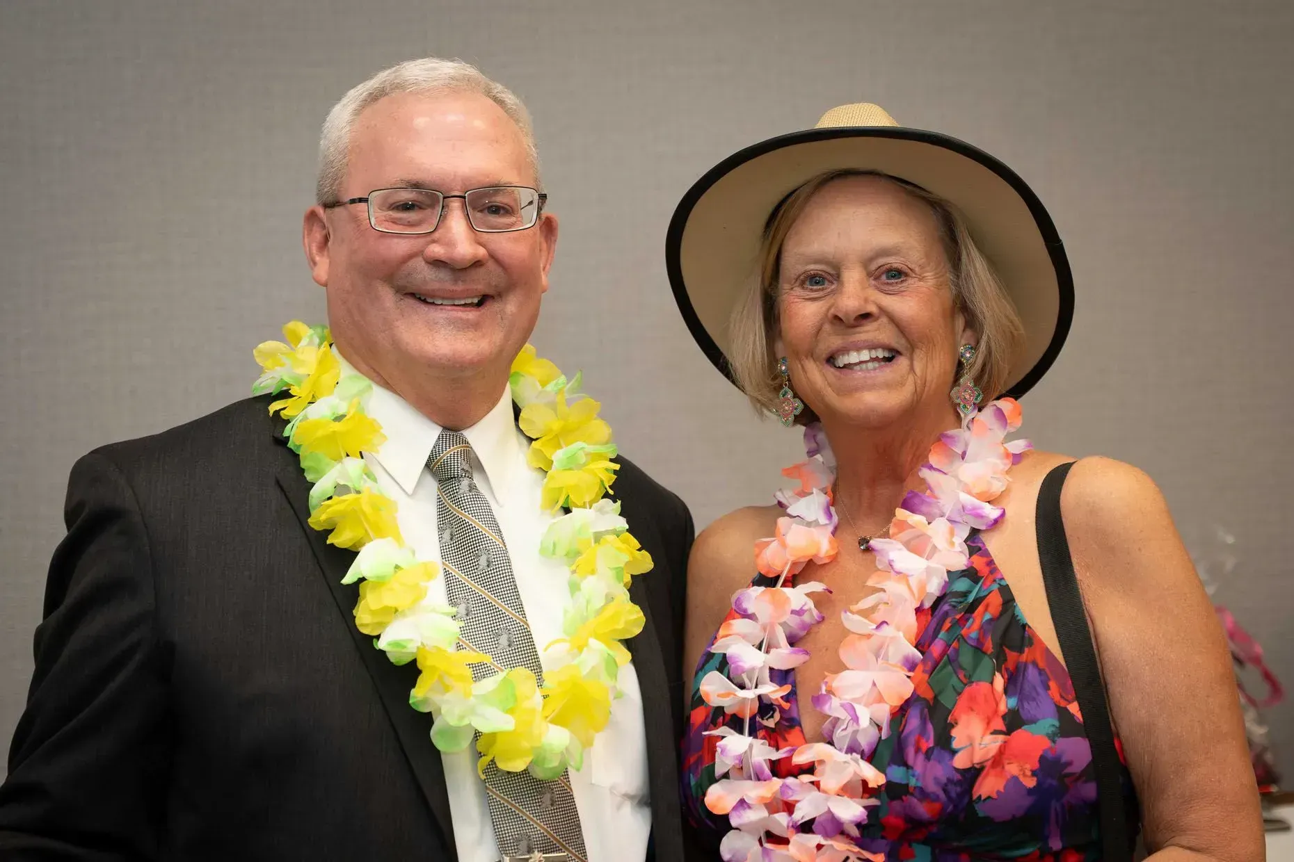 Man and woman smiling, wearing leis. Man in suit, woman in floral dress and hat.