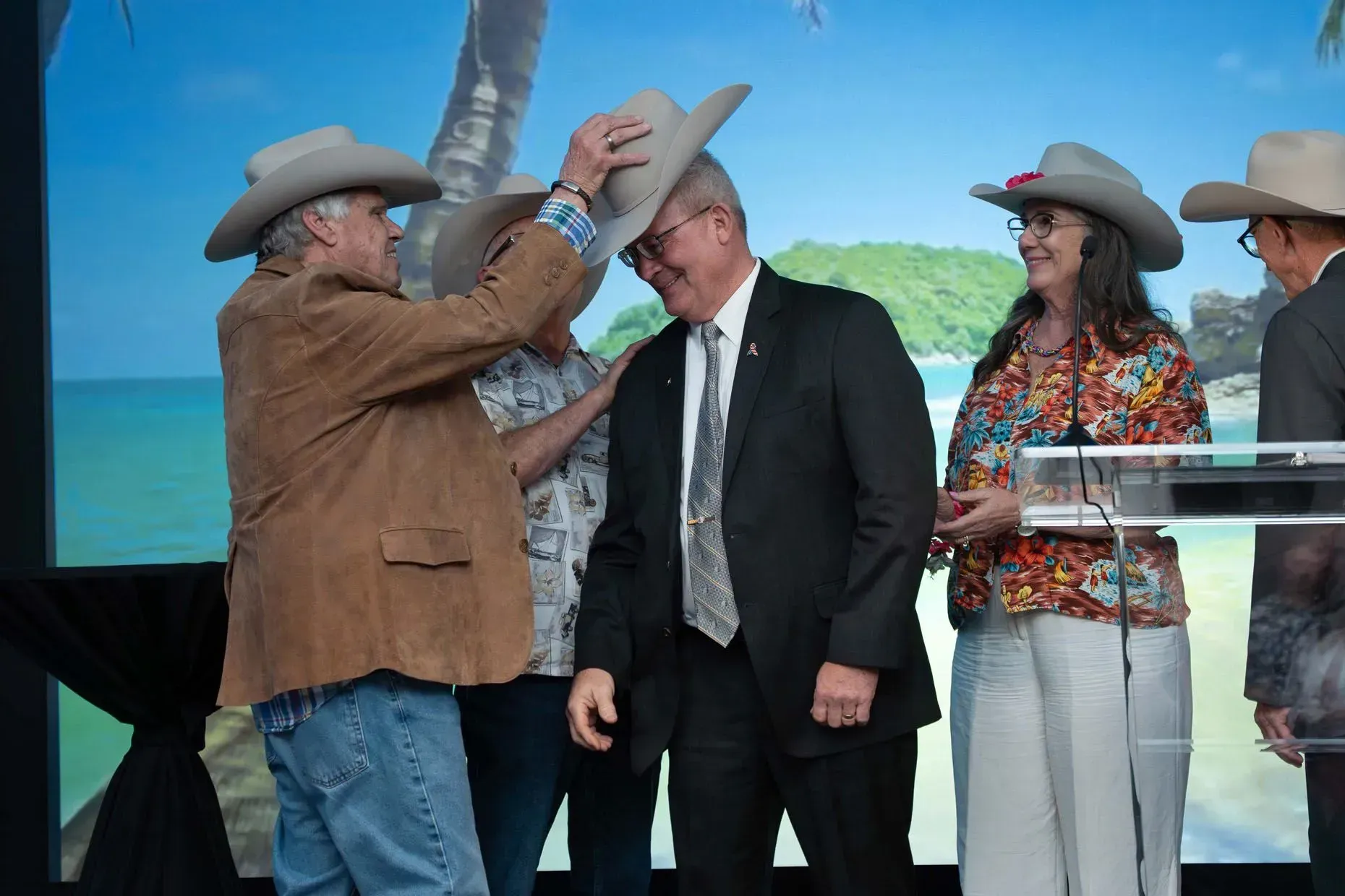 Man in suede jacket placing a cowboy hat on a man in a suit on stage, in front of a tropical backdrop.