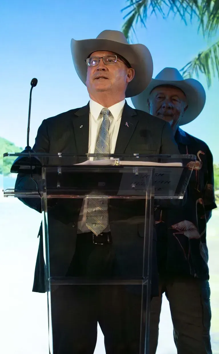 Man in cowboy hat speaking at podium, another man in background. Tropical scene backdrop.