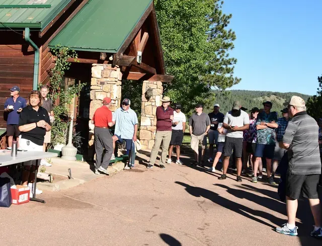 Group of people outdoors, gathered in front of a building with stone columns. Some are talking, others are listening.