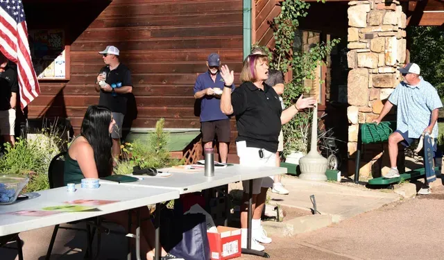 People at an outdoor event. Woman speaks from behind a table, others stand around. American flag visible.