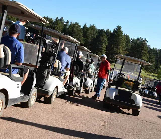 Golfers standing near golf carts lined up on a paved surface, preparing to play a round.