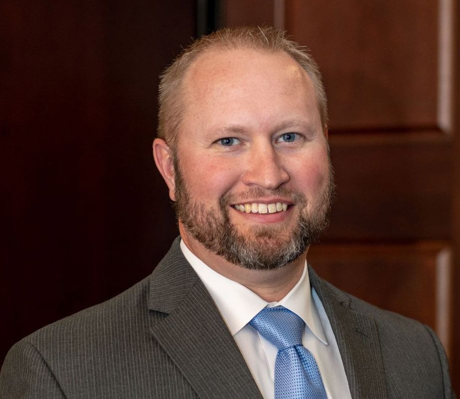 Man with a beard in a suit and tie smiles, indoors, against a brown background.