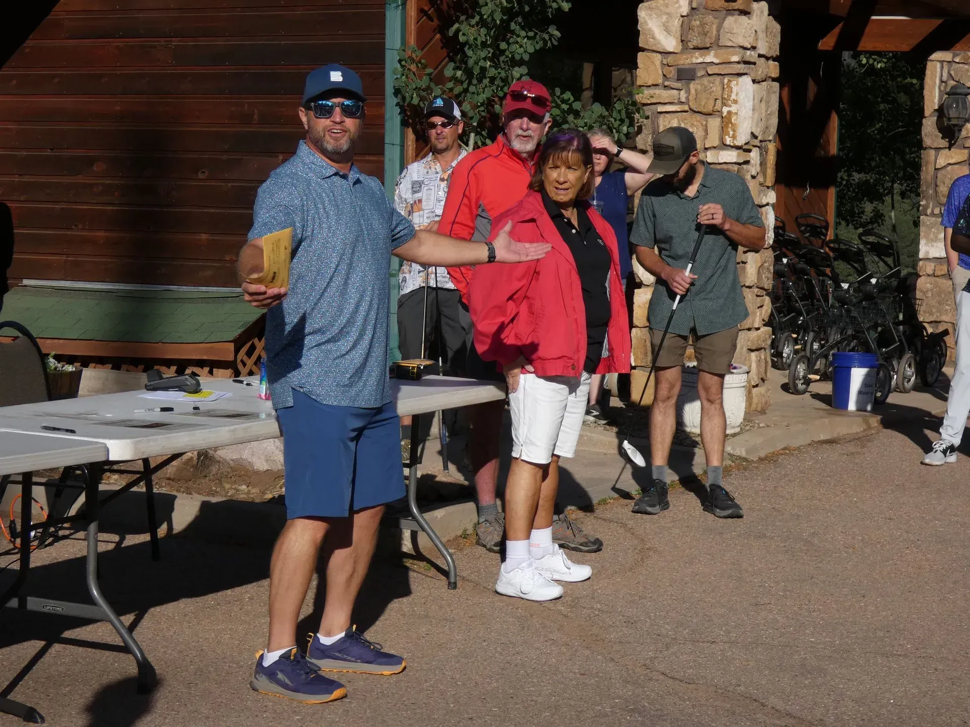 Group of people, some with golf clubs, at an outdoor event. A man in blue gestures.