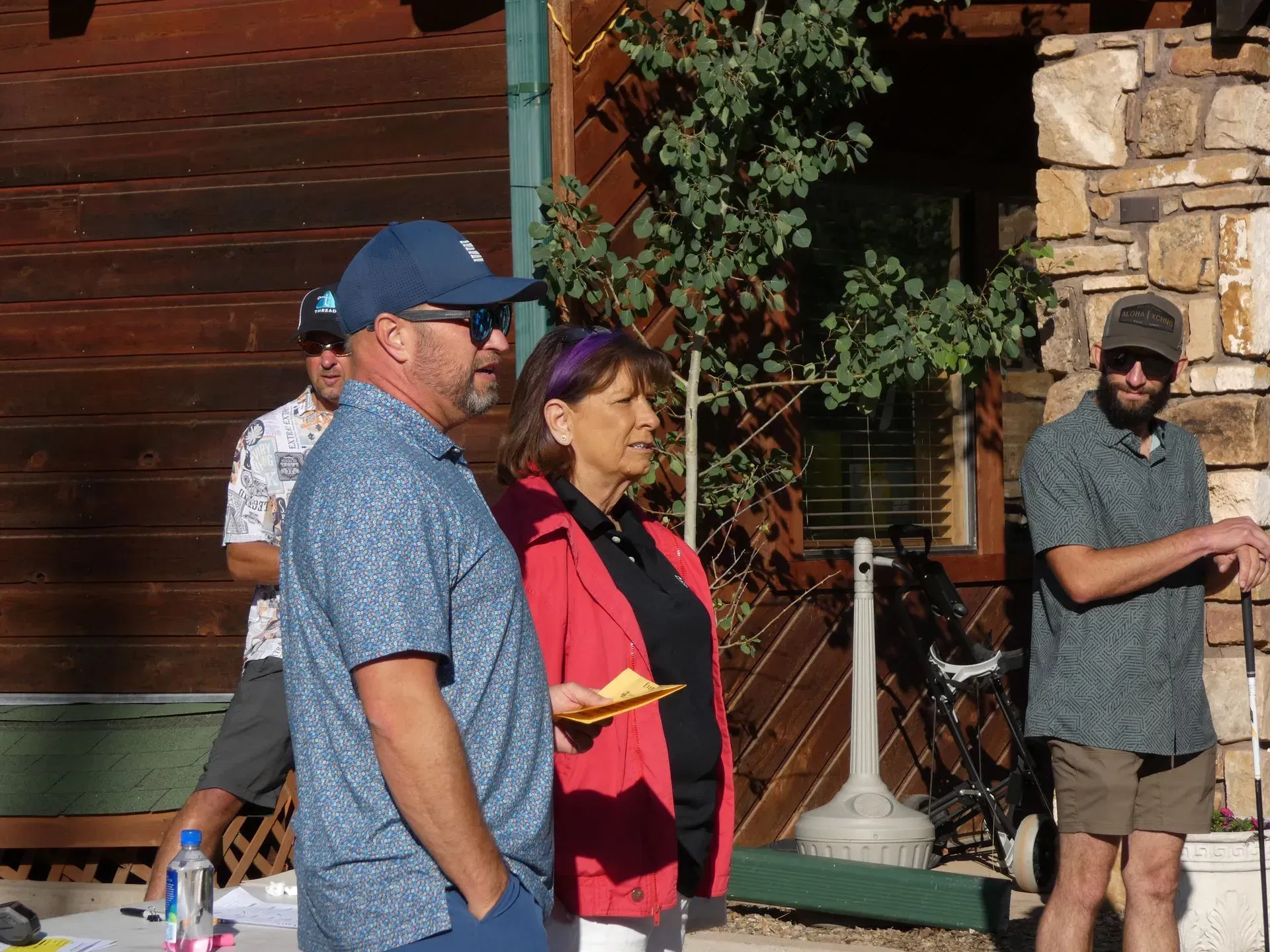 People at an outdoor gathering, some wearing sunglasses and casual clothing. Building in the background.