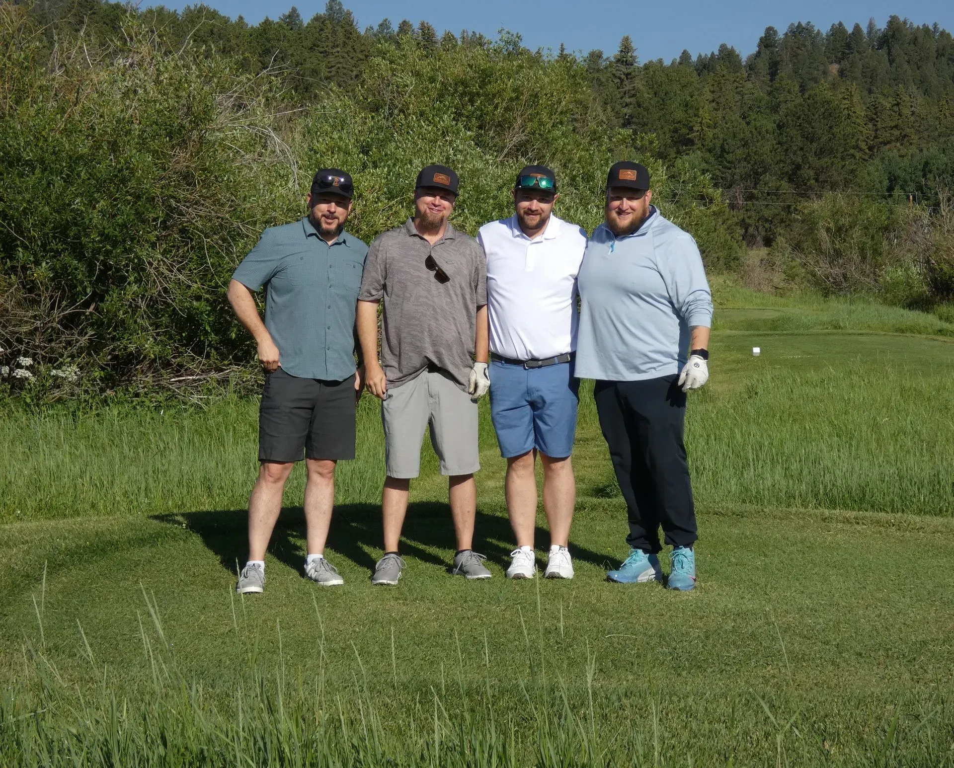 Four men stand together on a golf course. They wear casual attire, smiling, and posing for a photo.