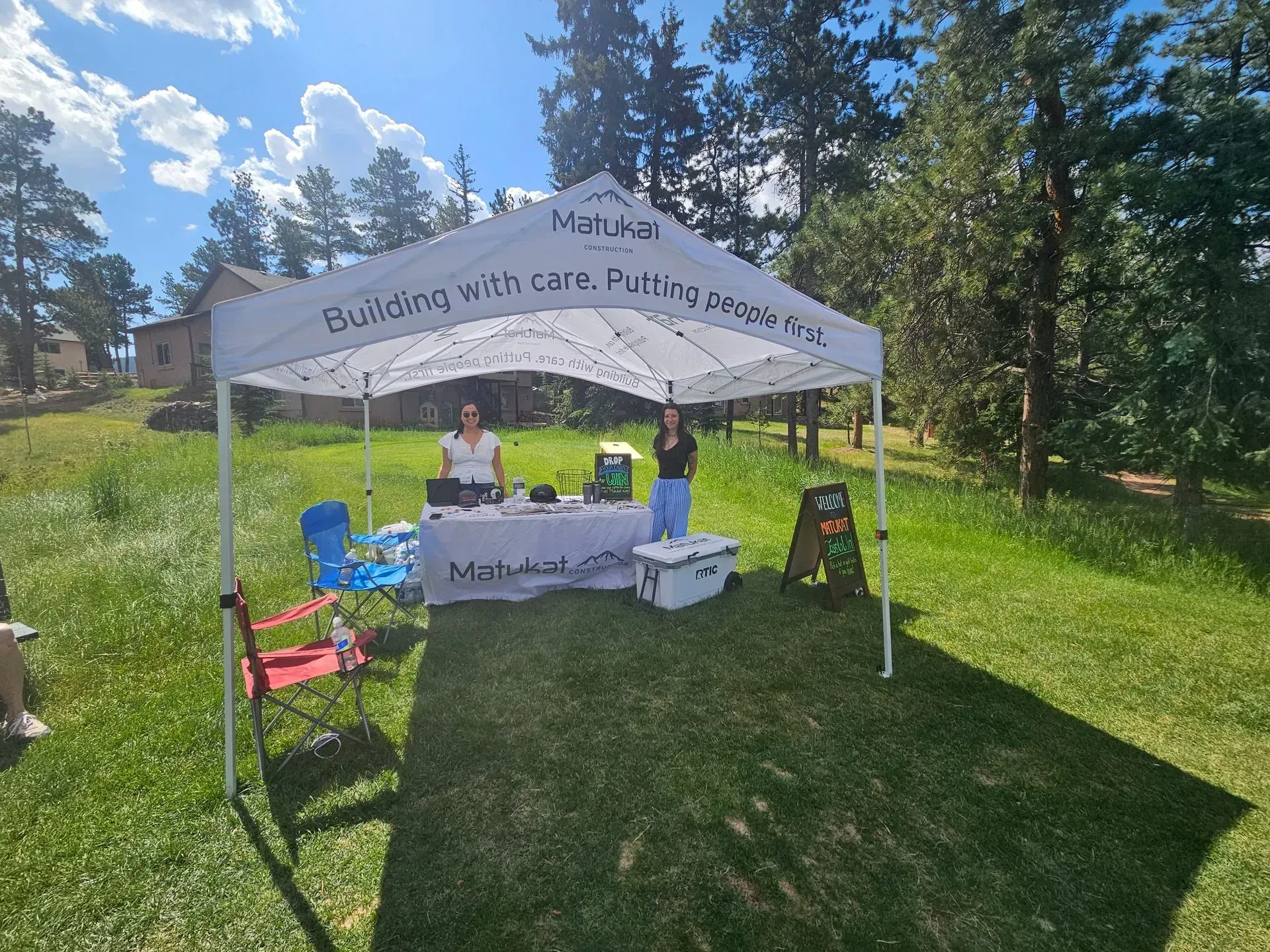 Two women at a white tent selling goods on a grassy hillside. Trees and a house are in the background.