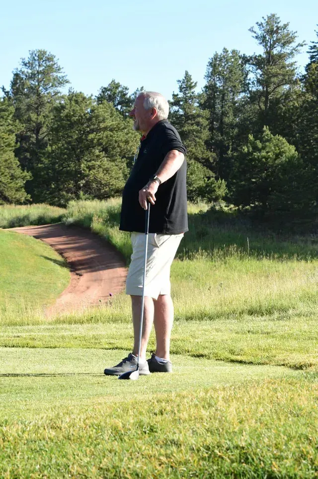 Man in black shirt and white shorts on a golf course, holding a golf club.