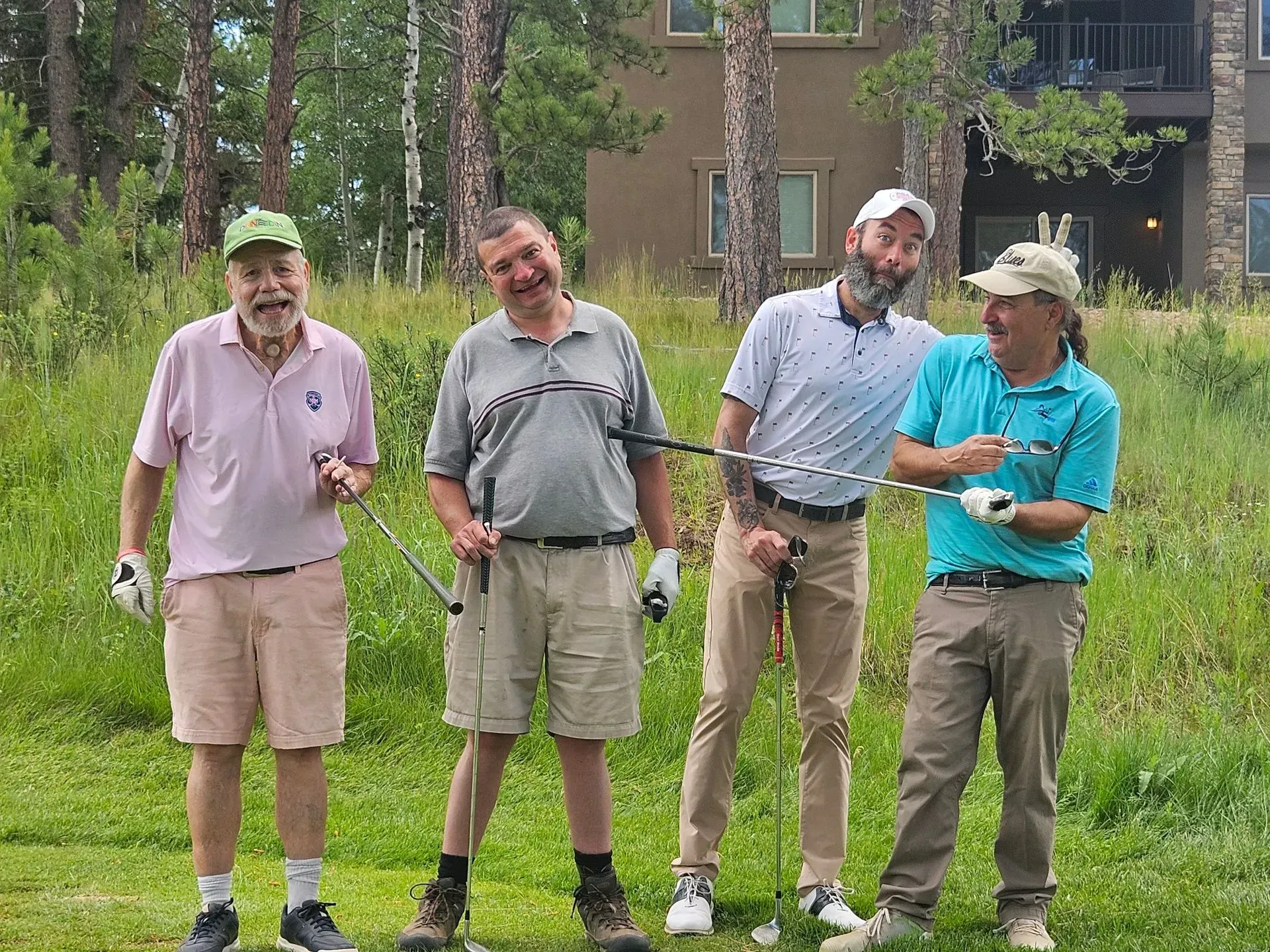 Four men on a golf course, holding clubs and smiling. Two are wearing hats. Green grass, trees, and a building in the background.