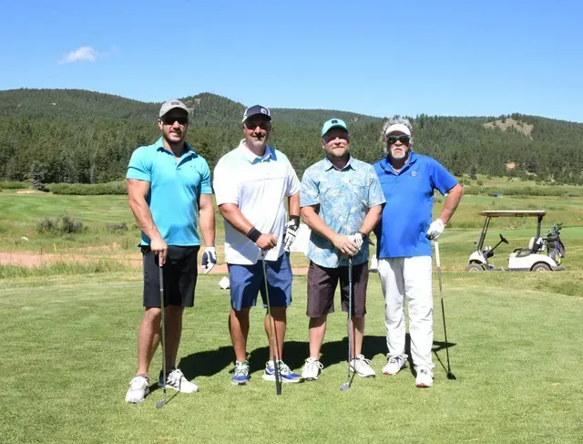 Four men pose on a golf course, holding clubs. Two wear blue, one white and floral, under blue sky, with hills in the background.