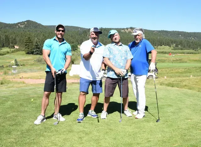 Four men on a golf course pose for a photo, smiling. Two wear shorts, two wear white pants, blue and white shirts.