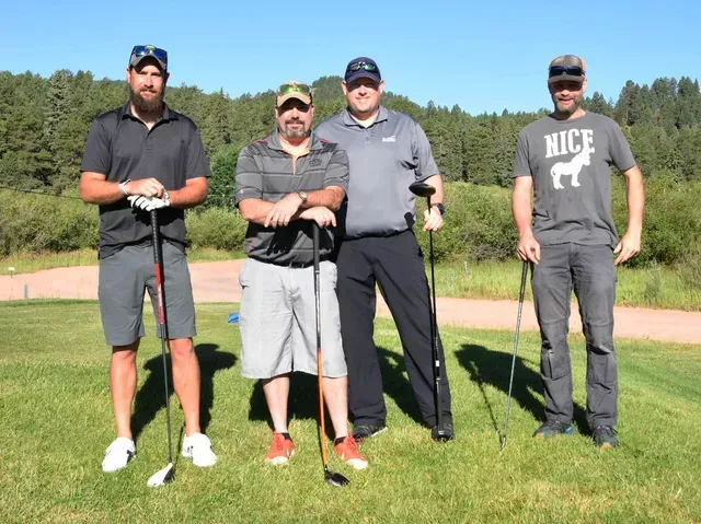 Four men posing on a golf course, holding clubs. One is using crutches. Green grass, sunny day.