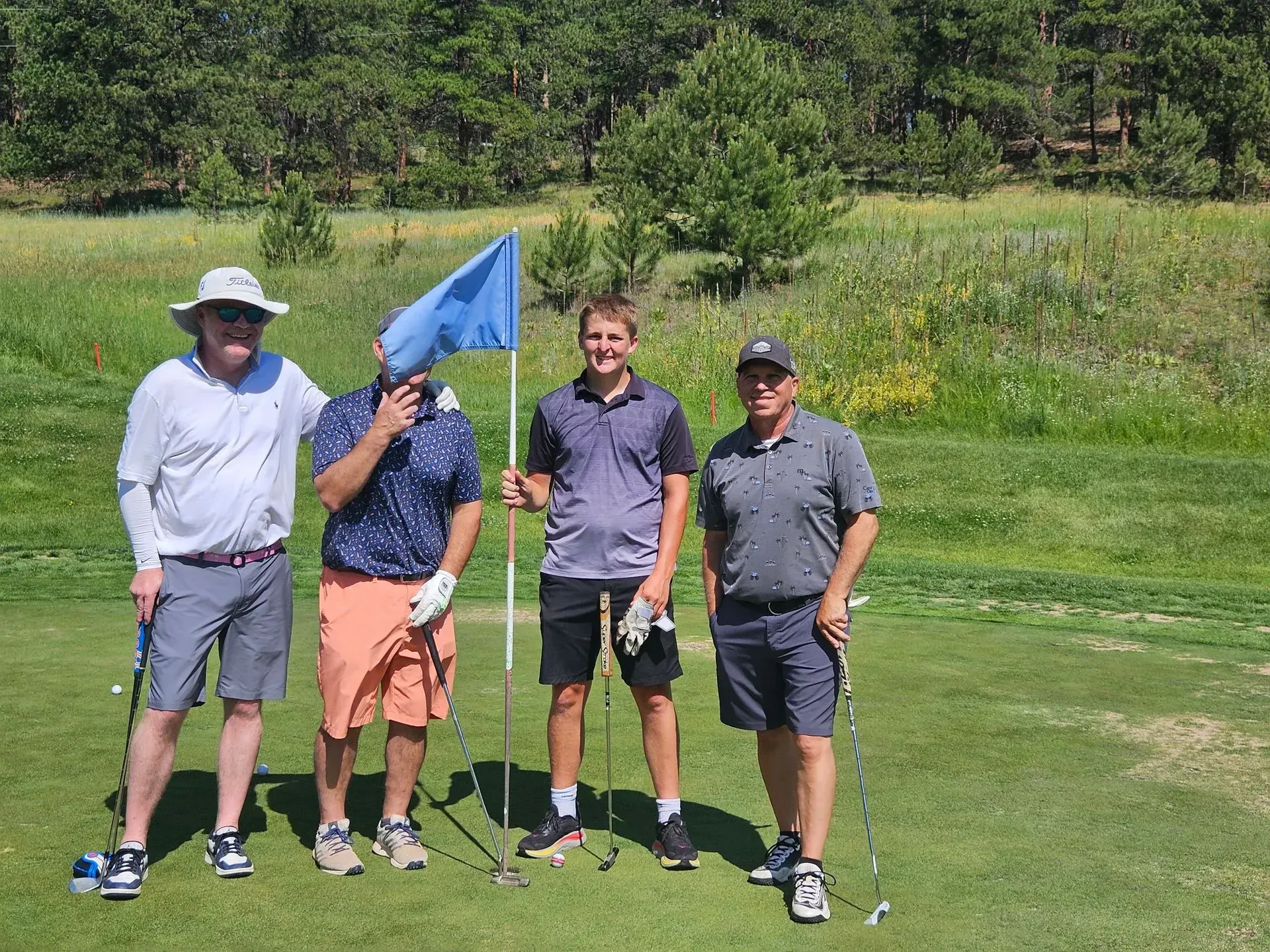 Four golfers on a green, smiling, holding clubs and a flag. Sunny day, trees and grass in background.