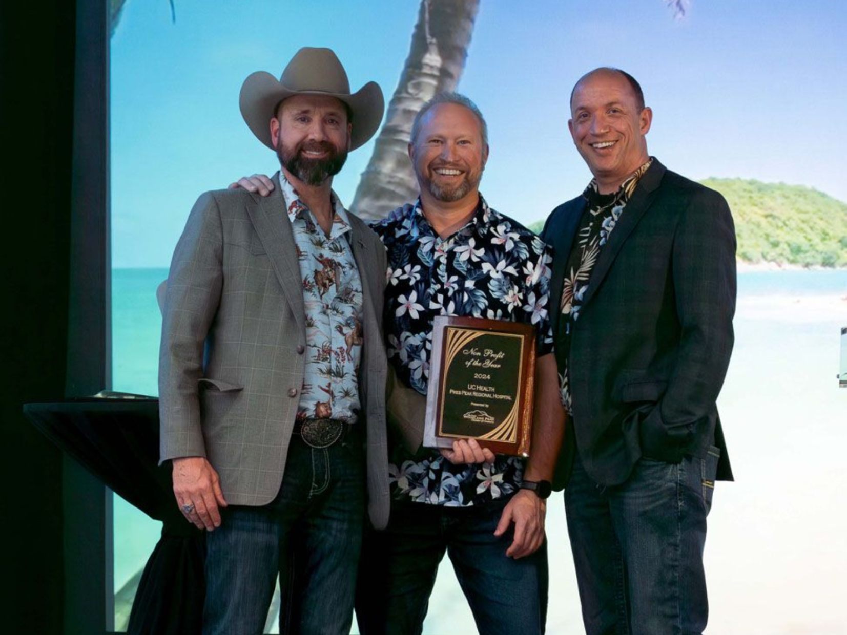 Four people on stage at a podium. Woman in beige jacket, man with beard in blazer, others smile.
