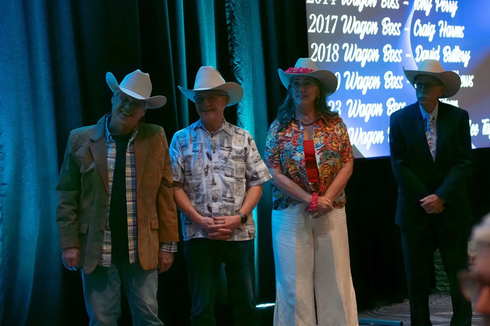 Four people in cowboy hats on stage; two men and one woman smiling, one man with a neutral expression.
