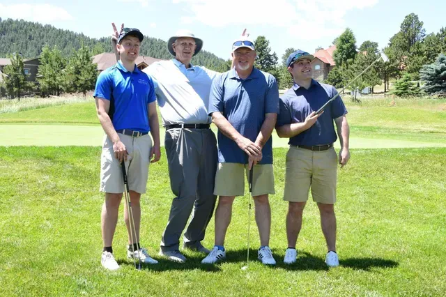 Four men on a golf course, posing for photo. One wearing a headdress. Sunny day, green grass, mountains in the background.