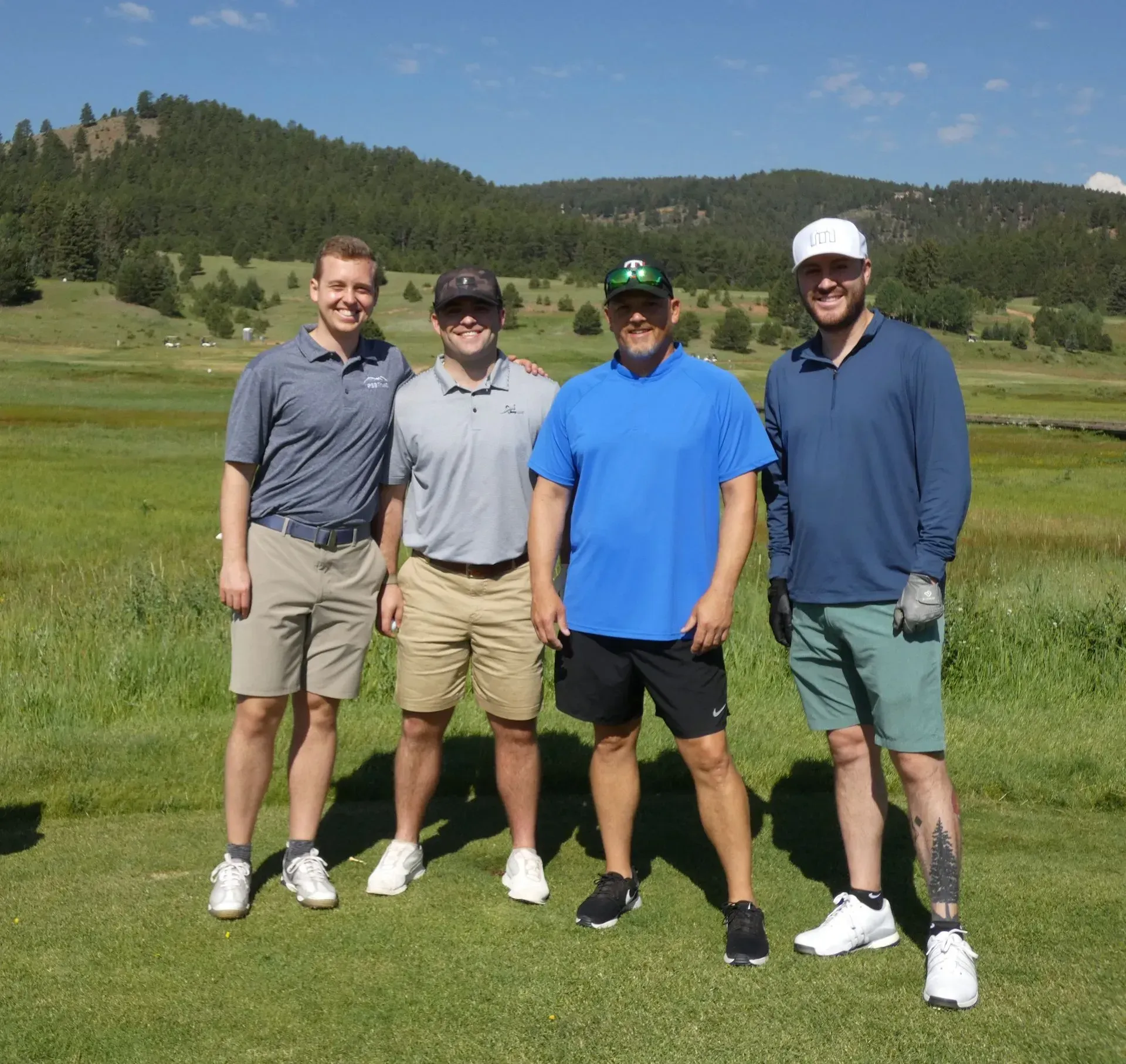 Four men pose on a sunny golf course; mountains in the background.