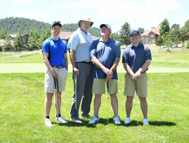 Four men in golf attire on a green, two smiling, mountain backdrop.