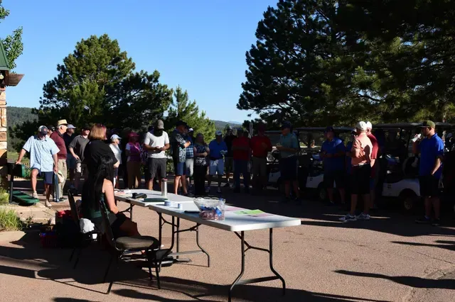 People gathered near a table outside; golf carts nearby. Blue sky, trees in the background.