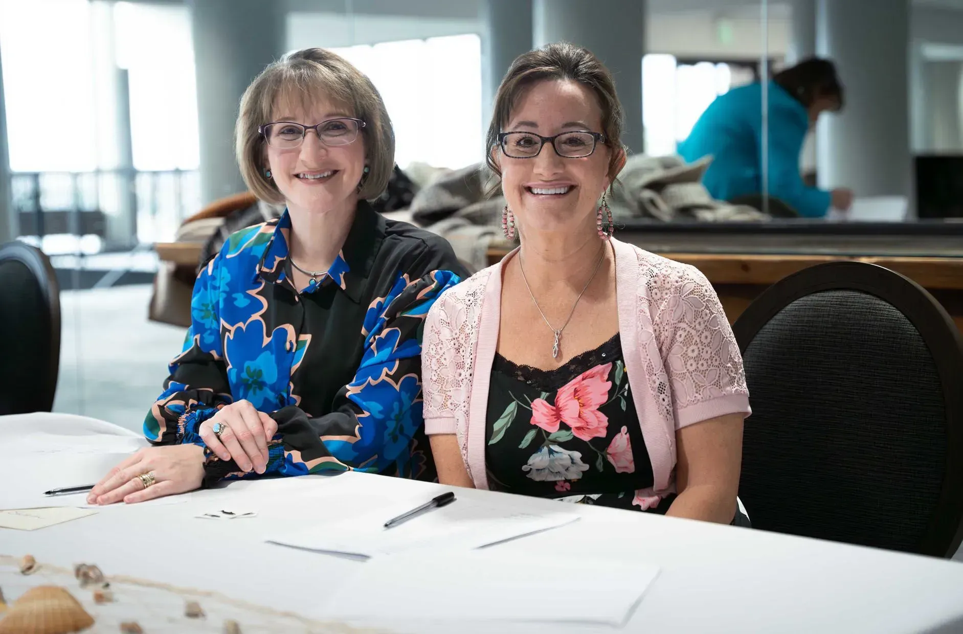 Two women seated at a table smile. One wears a blue floral jacket, the other a pink cardigan over a floral top.