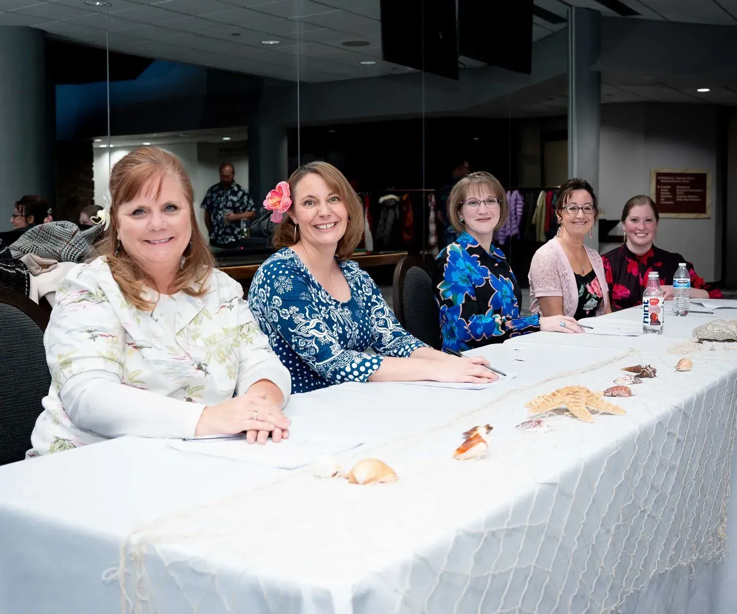 Five women smiling at a table with shells. Indoor setting with a white tablecloth.