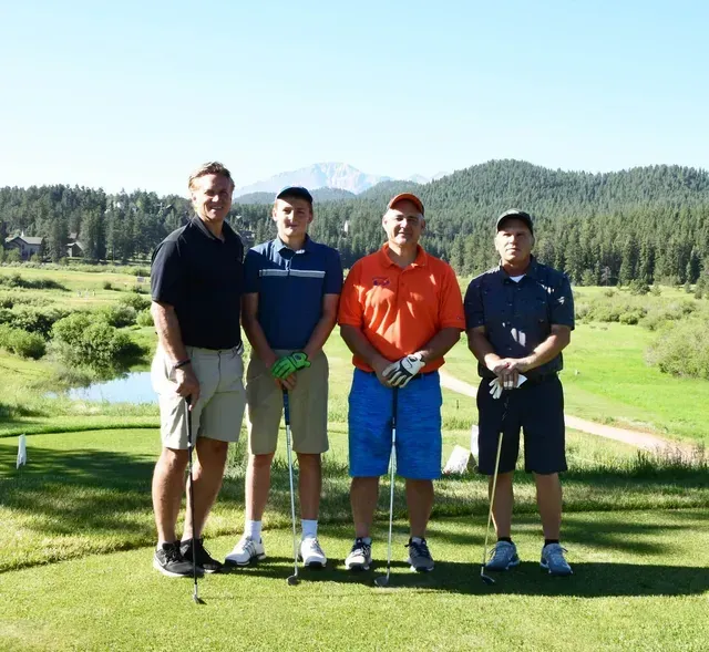 Four men standing on a golf course, posing for a photo. Green grass, trees, and mountains in the background.