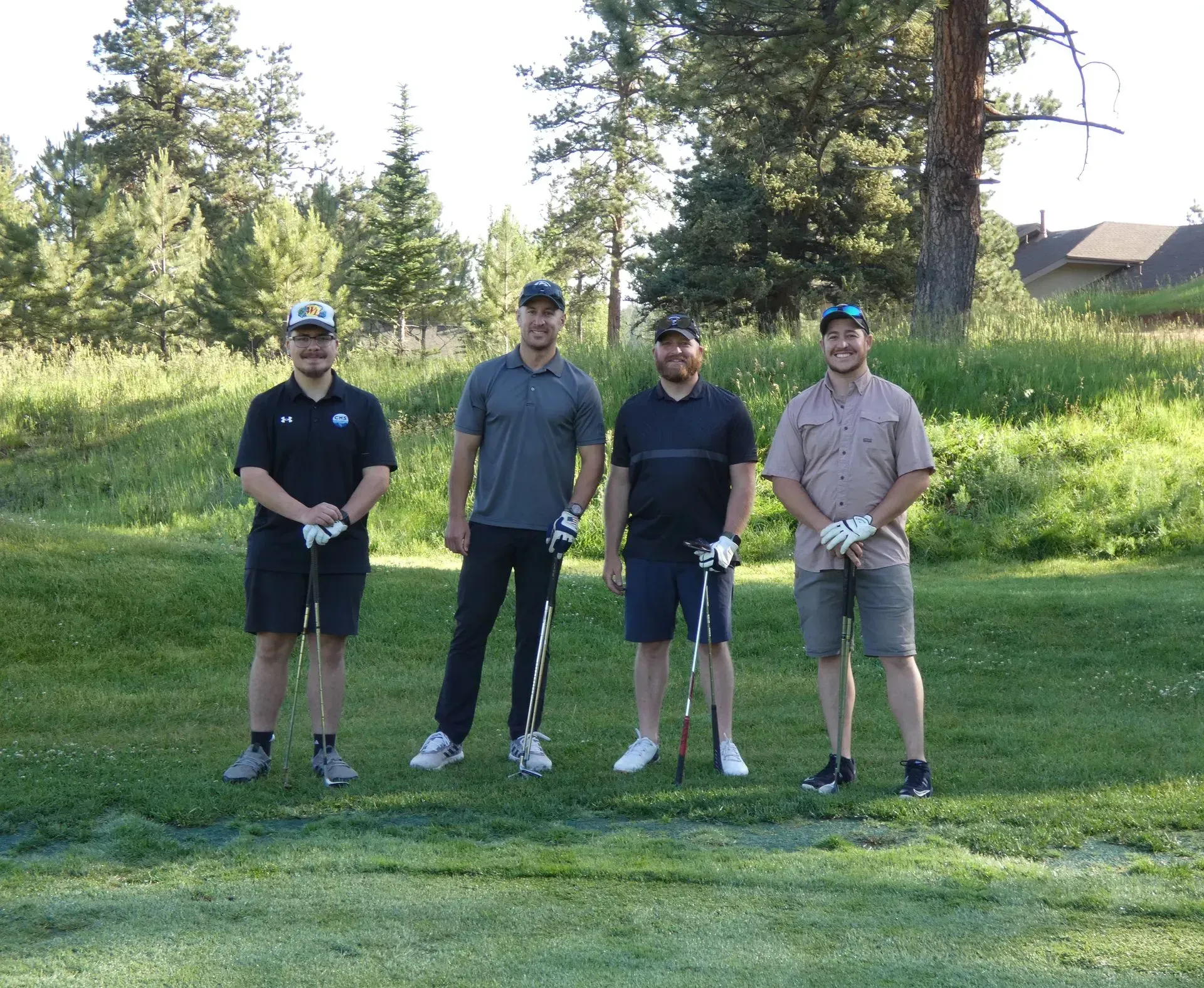 Four men standing on a golf course, holding golf clubs, smiling. Sunny day, green grass, trees in the background.