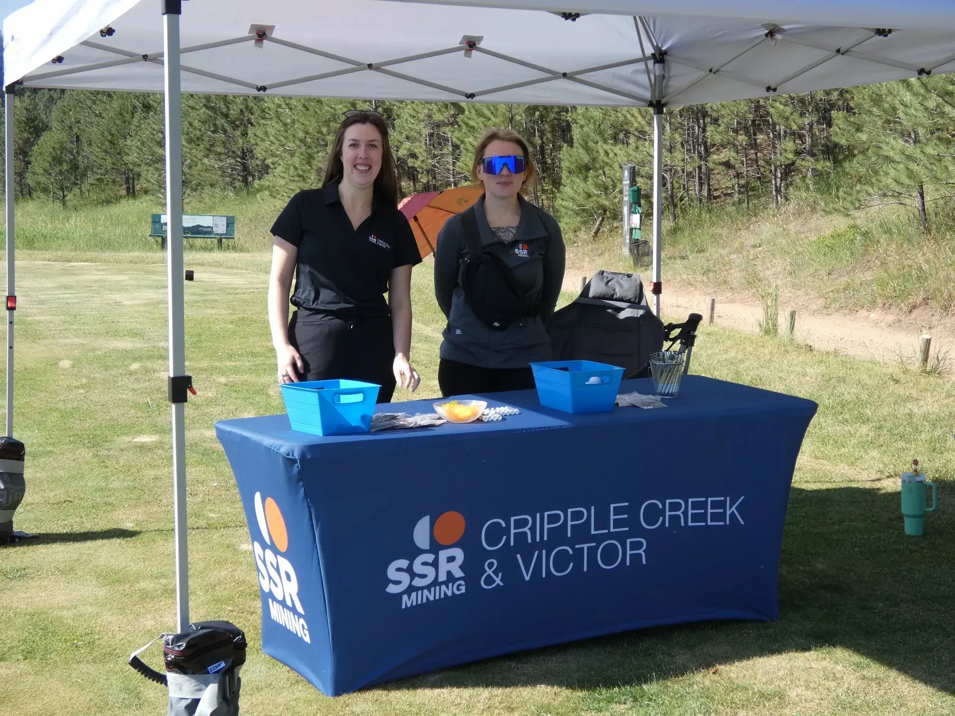 Two women at a Cripple Creek & Victor booth under a canopy; blue table cover, clear sky.