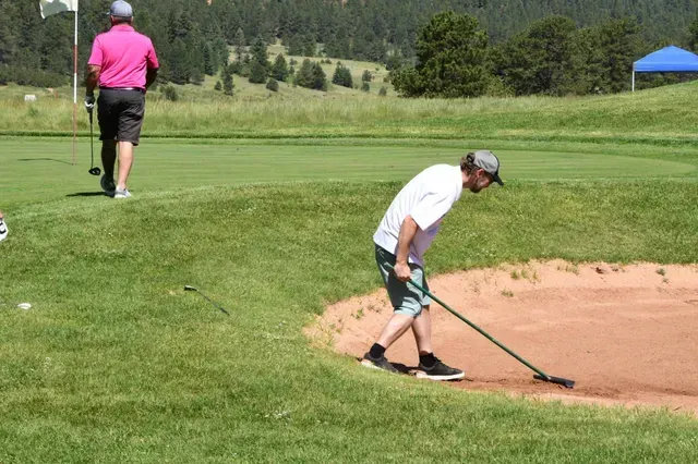 Man raking sand trap on golf course; another golfer walks on green.