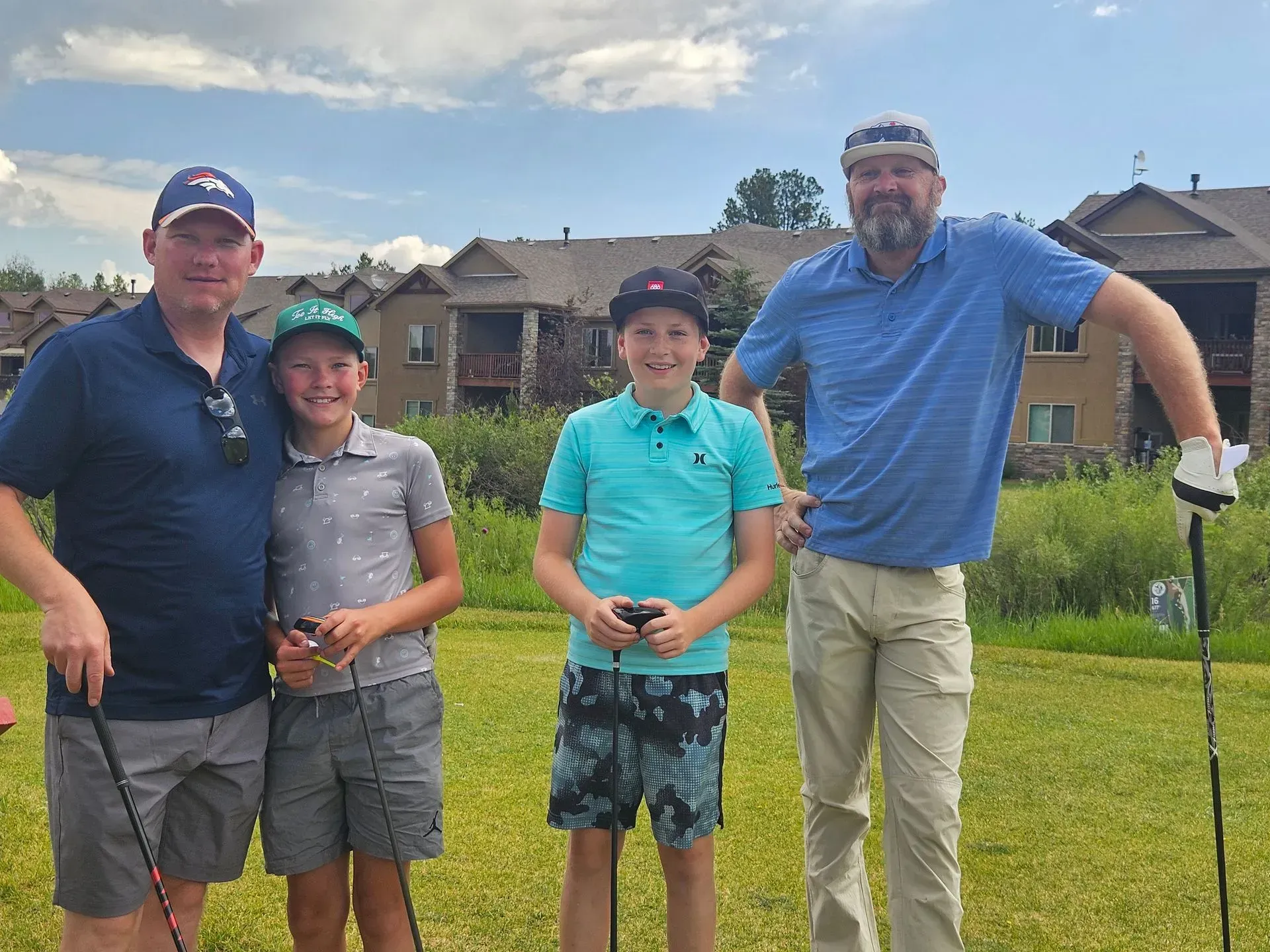 Four people smiling on a golf course; two adults, two children. Green grass, blue sky, buildings in background.