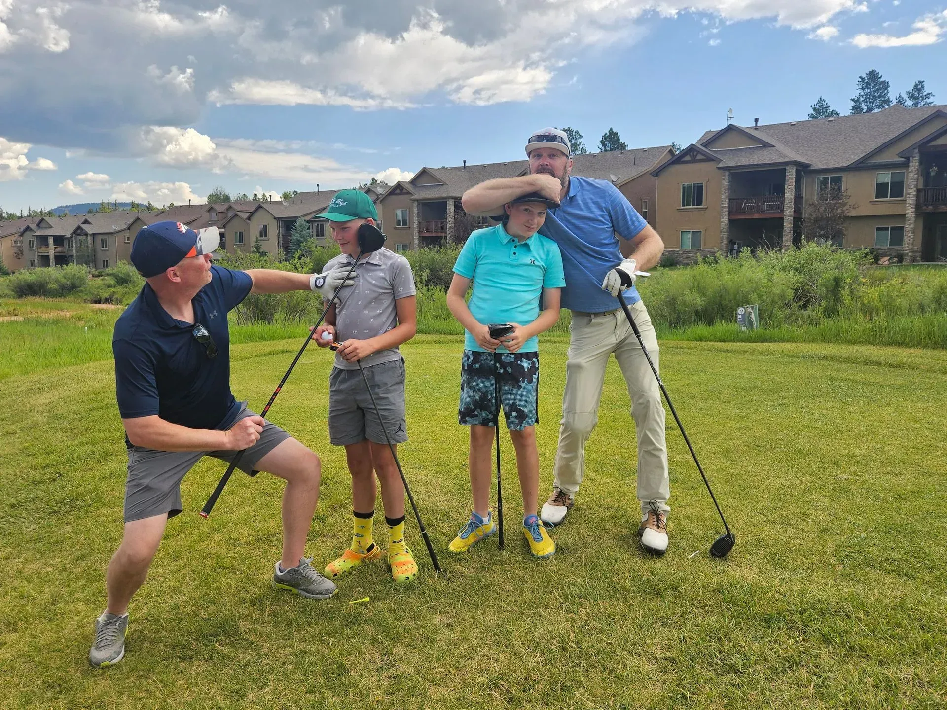 Four people on a golf course; two men and two boys. They are posing playfully with golf clubs.
