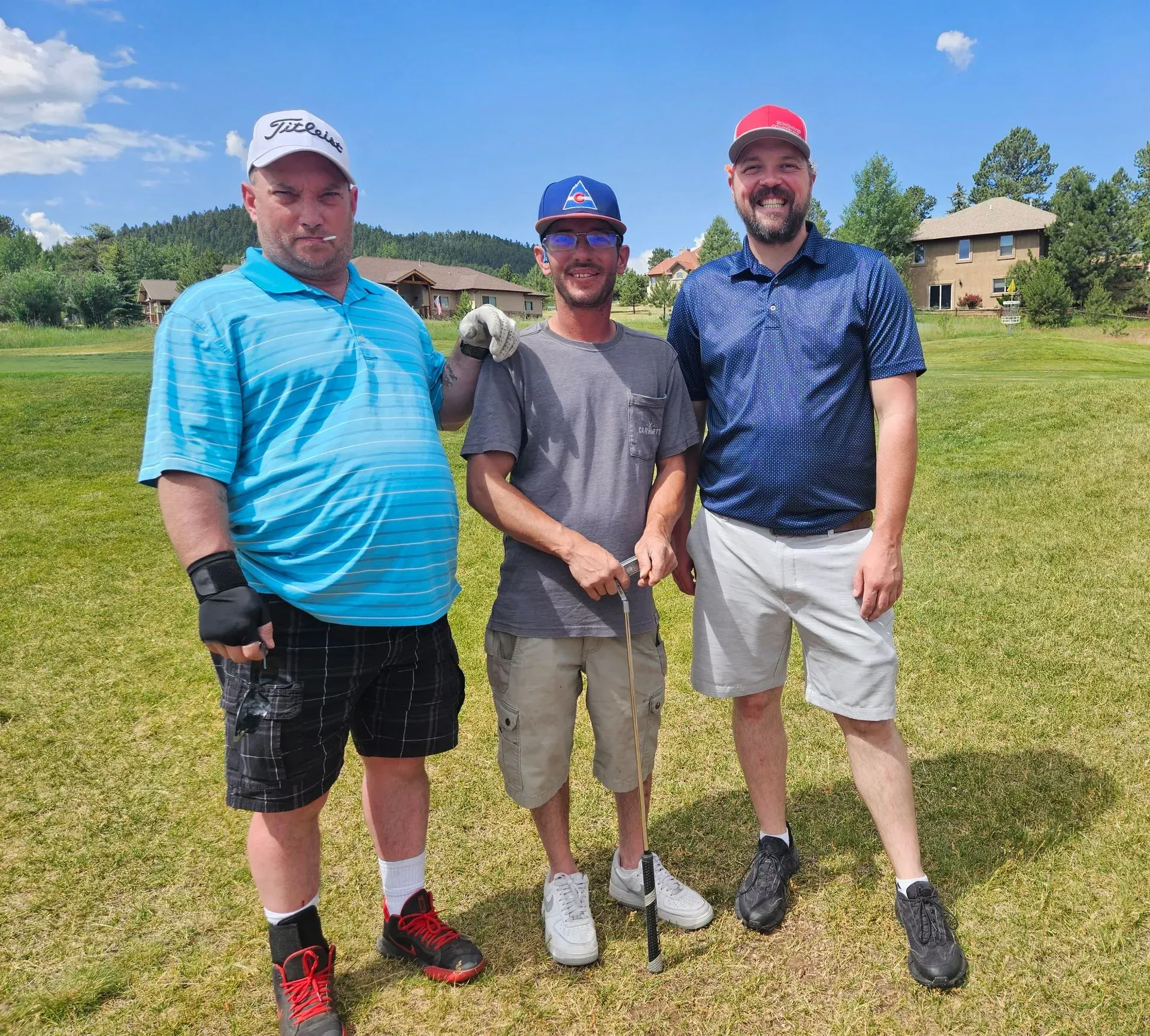 Three men pose on a golf course, smiling. One wears a red cap, another a blue shirt and shorts, the third, a turquoise polo.