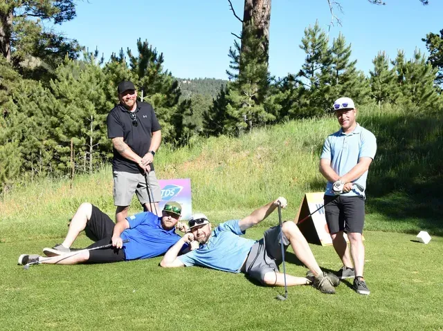 Four men pose on a golf course: two lying down, two standing with clubs, sunny day.