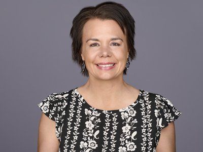 Woman with short brown hair, smiling at the camera; wearing a black and white floral print top against a gray backdrop.