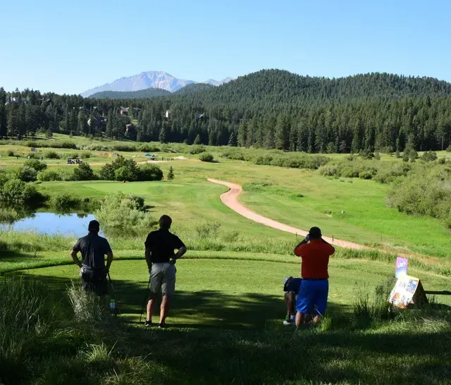 Golfers on a tee box, overlooking a green course with a mountain backdrop.