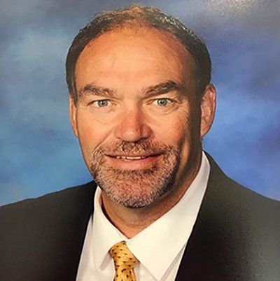 Man in suit smiling, with short brown hair and beard. Blue background.