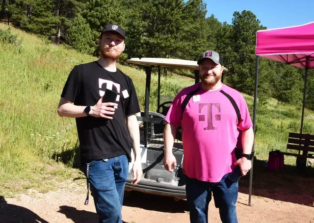 Two men in T-Mobile shirts stand next to a golf cart and pink tent outdoors. One man in black gives a thumbs up.