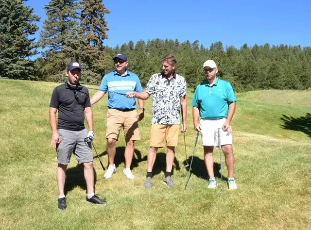 Four men on a golf course, one pointing, smiling. Green grass, trees, blue sky.