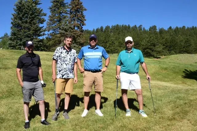 Four men on a golf course, sunny day. Men in shorts and golf attire, standing in the grass holding golf clubs.