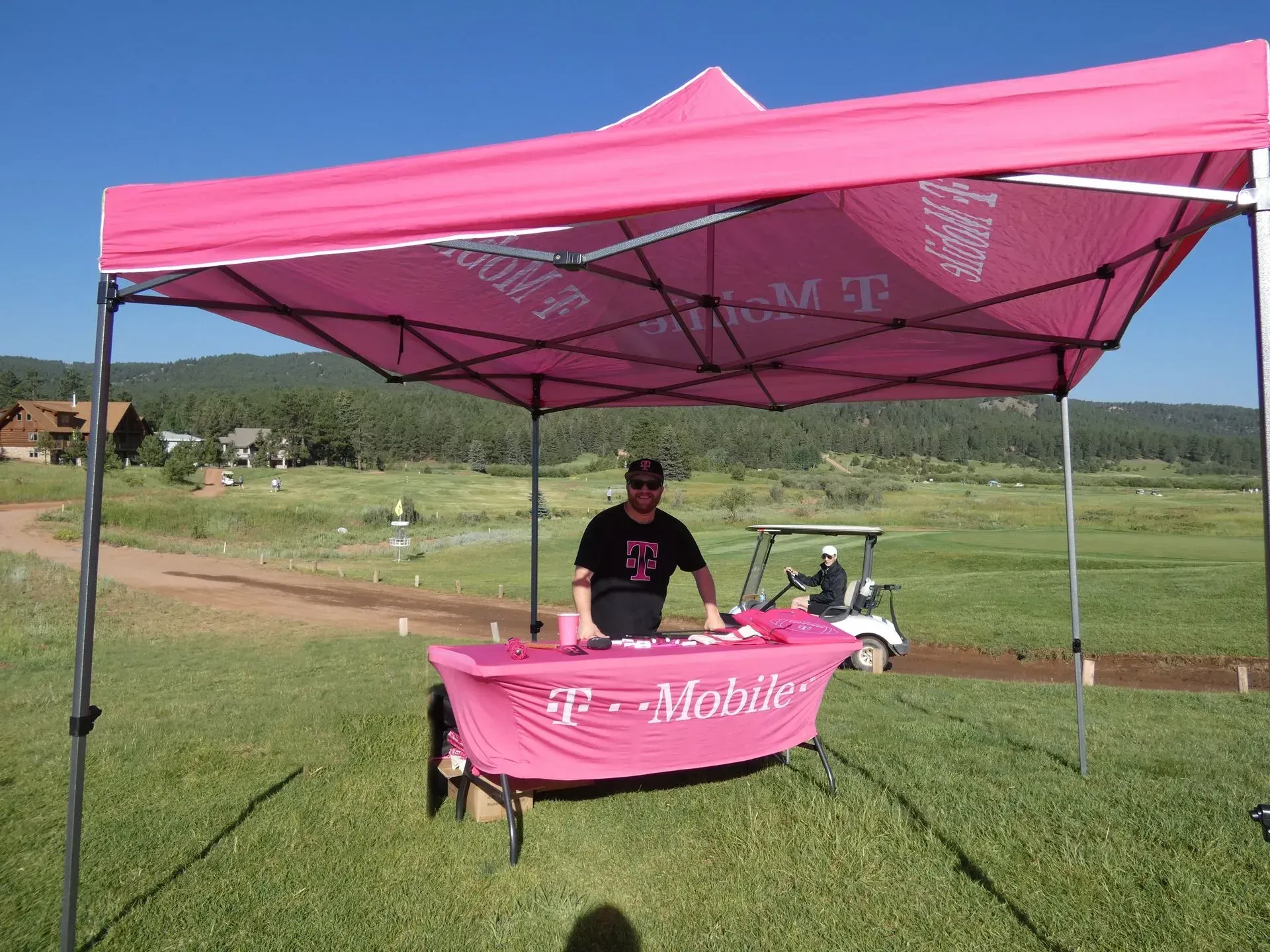 Person under a pink T-Mobile tent at a golf course, manning a branded table, golf cart in background.