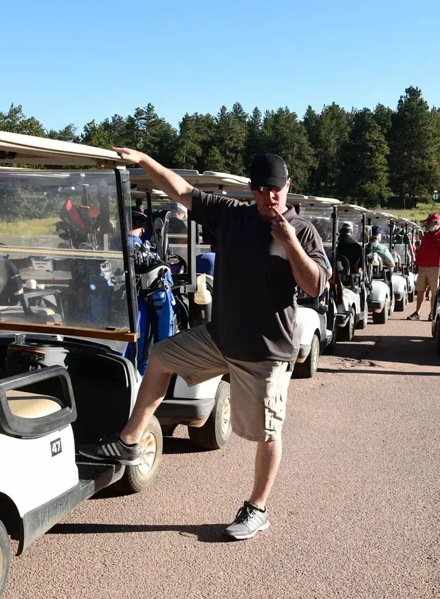 Man in hat and shorts poses by golf cart, holding snack, other carts in background, trees, sunny day.