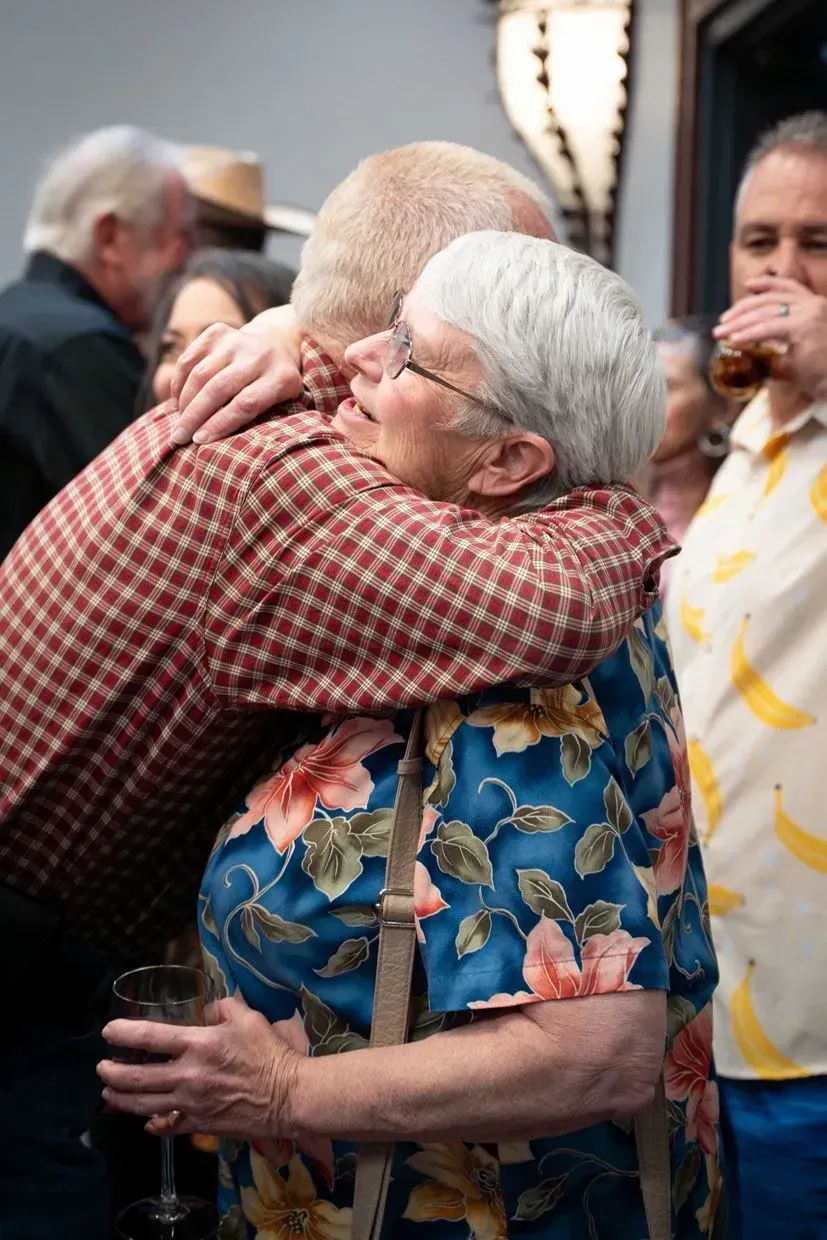Two older adults embracing outdoors; woman in floral shirt, man in plaid. Other people in the background.