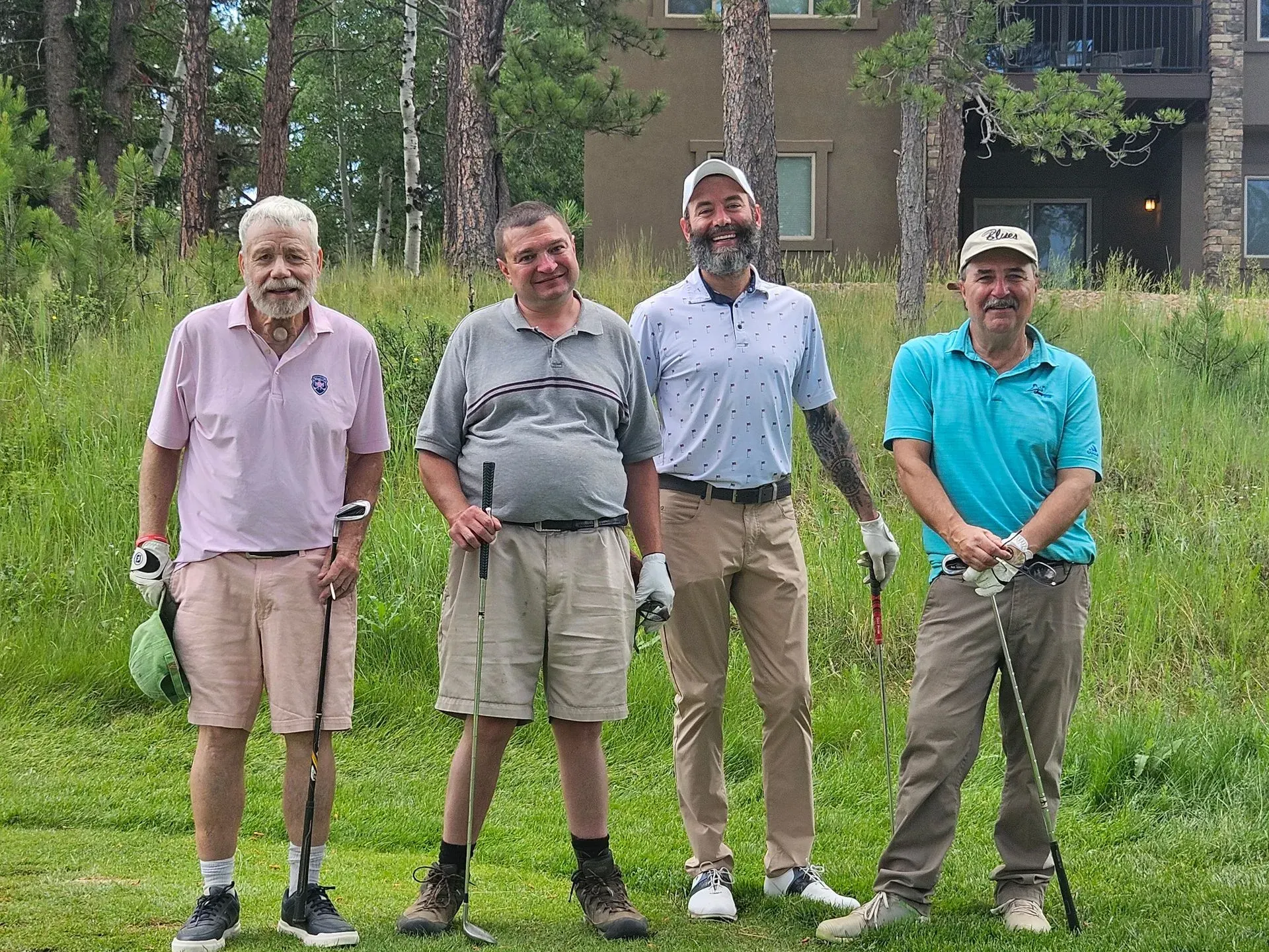 Four men in golf attire standing on a grassy course, smiling and holding clubs. Trees and a building are in the background.