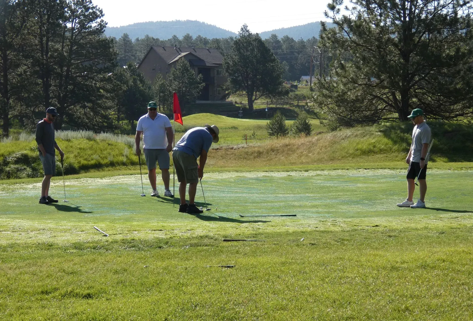 Golfers on a putting green; one lines up a putt, others watch, red flag in hole, mountain backdrop.