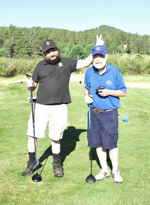 Two men on a golf course. One makes a bunny ear gesture on the otherās head. Sunny day.