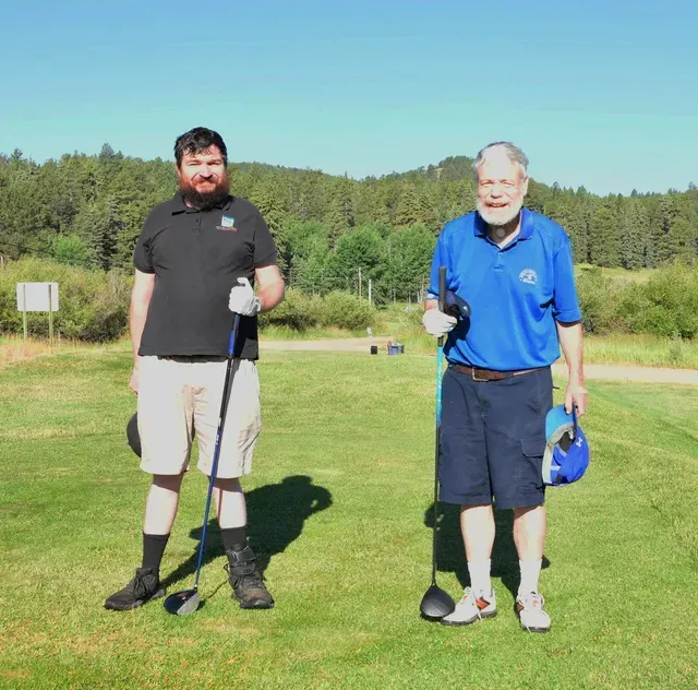 Two men on a golf course: one with beard in black shirt, the other older, in blue shirt.